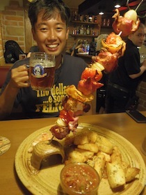 大きな肉の串焼きを前にビールを片手に持った男性